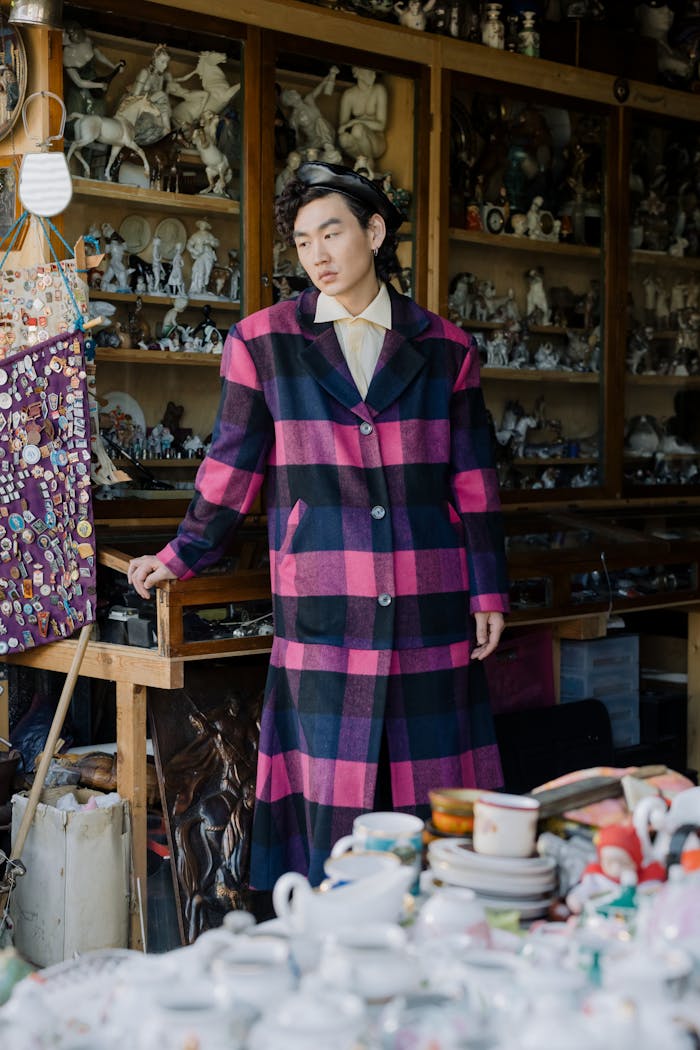An adult male fashionably dressed in a checkered coat poses inside a curio shop filled with porcelain figurines.